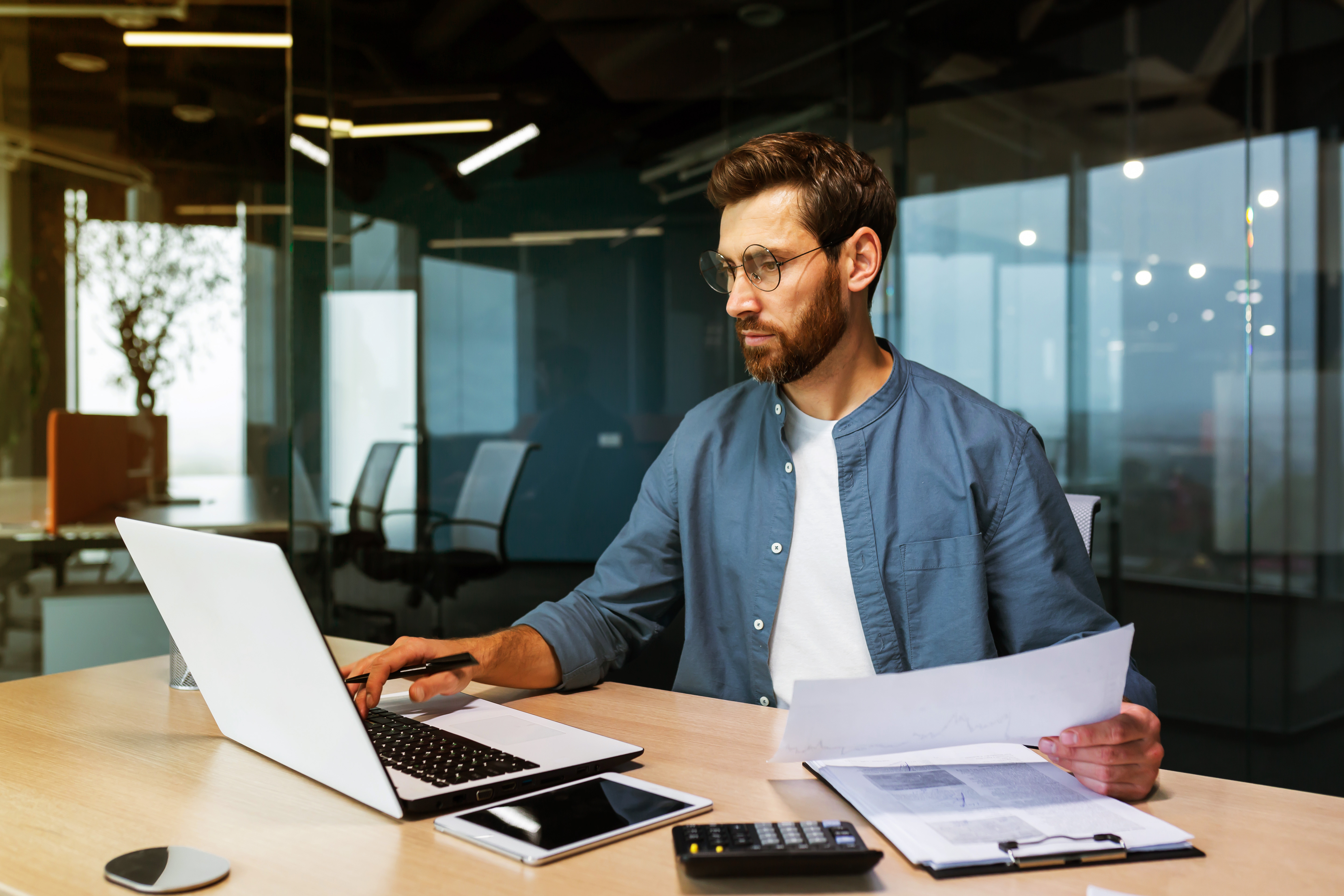 Man with glasses working on laptop and holding papers in a modern office setting