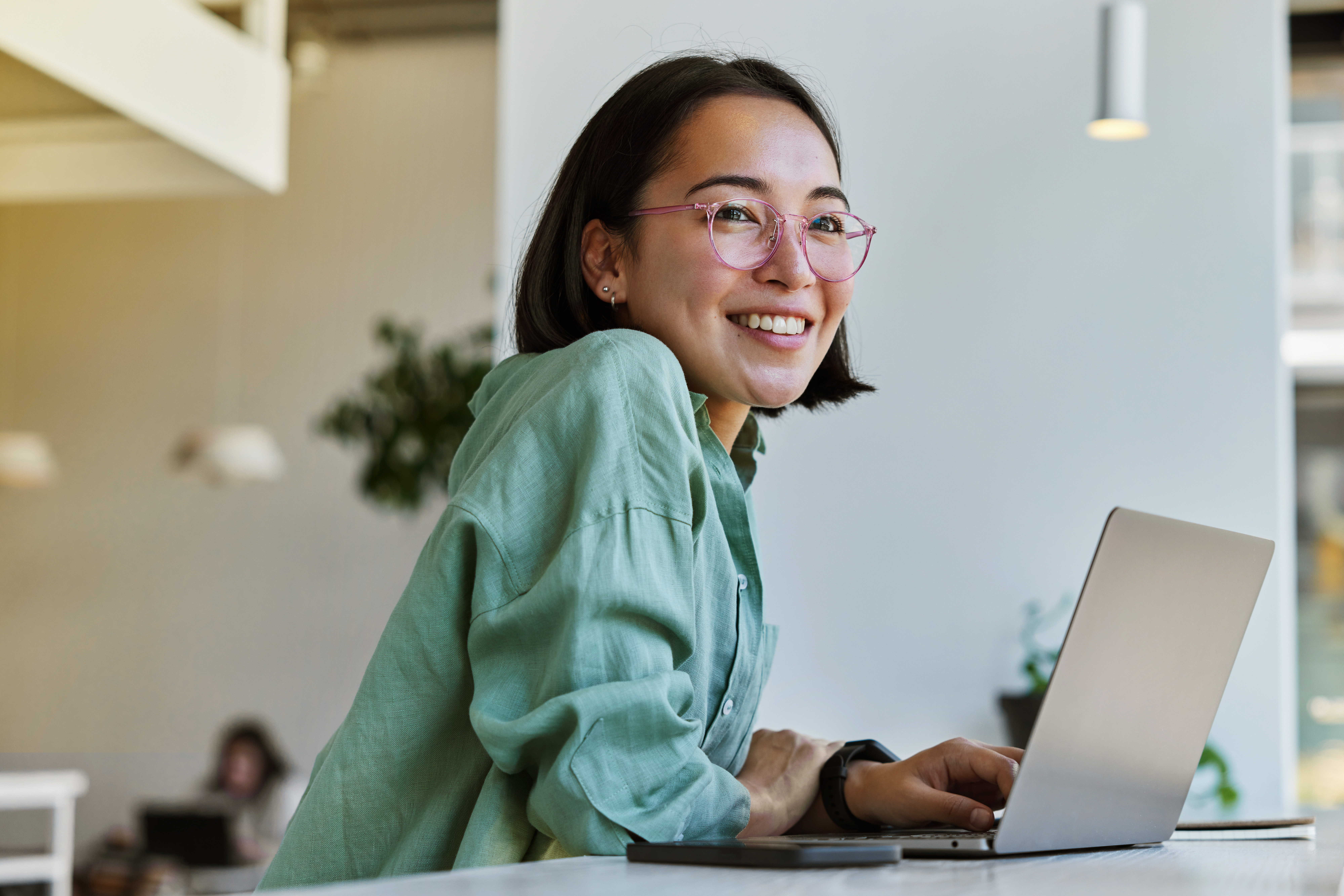 Woman with glasses and green shirt smiling while using a laptop at a cafe