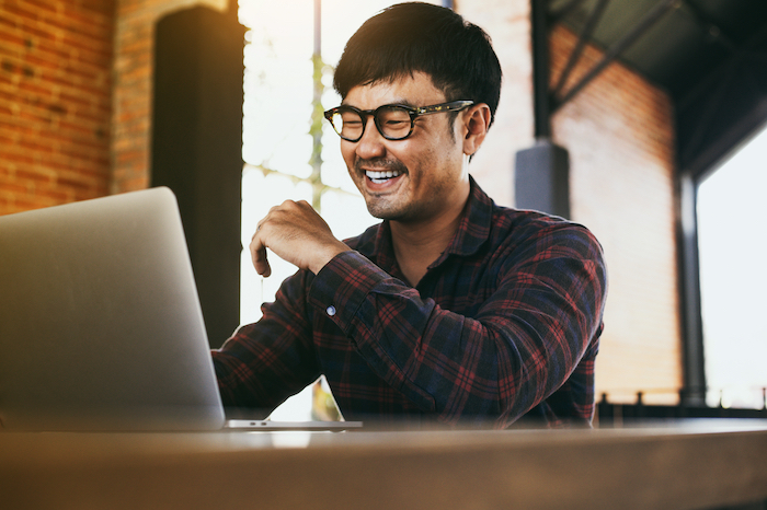 Smiling man wearing glasses and a plaid shirt using a laptop in a bright, modern cafe.