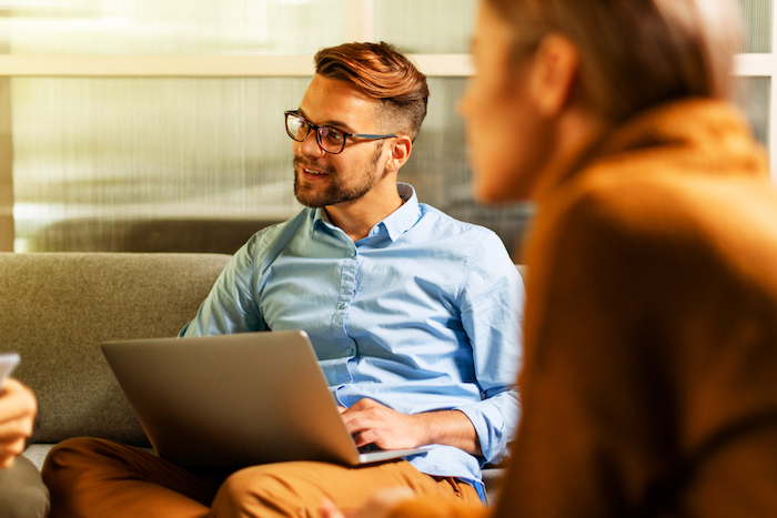 Man with glasses using a laptop while talking with a female colleague in an office setting.