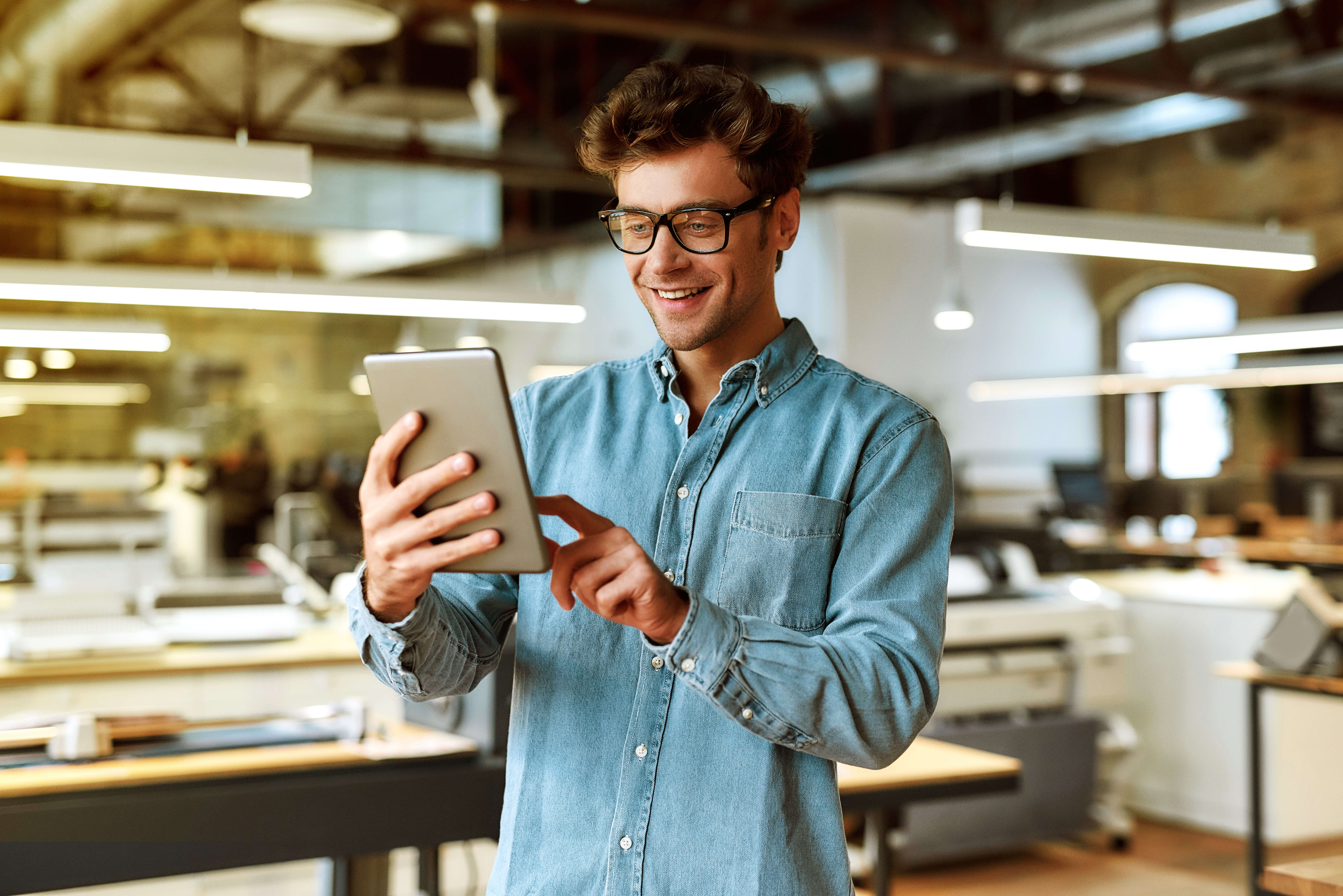 Man with glasses in denim shirt using a tablet in a modern office setting.