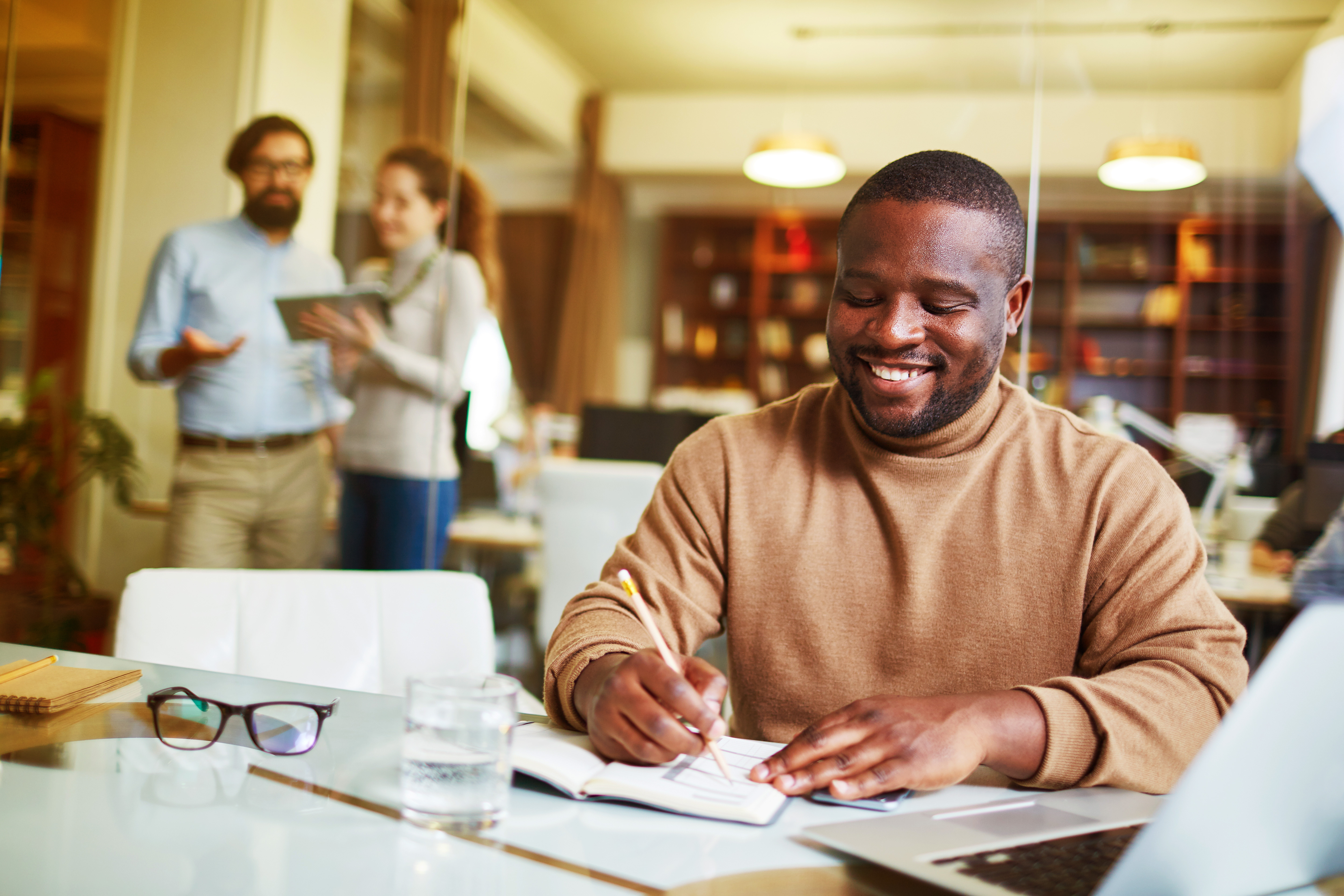 Man smiling and writing in notebook at office desk, with colleagues talking in the background.
