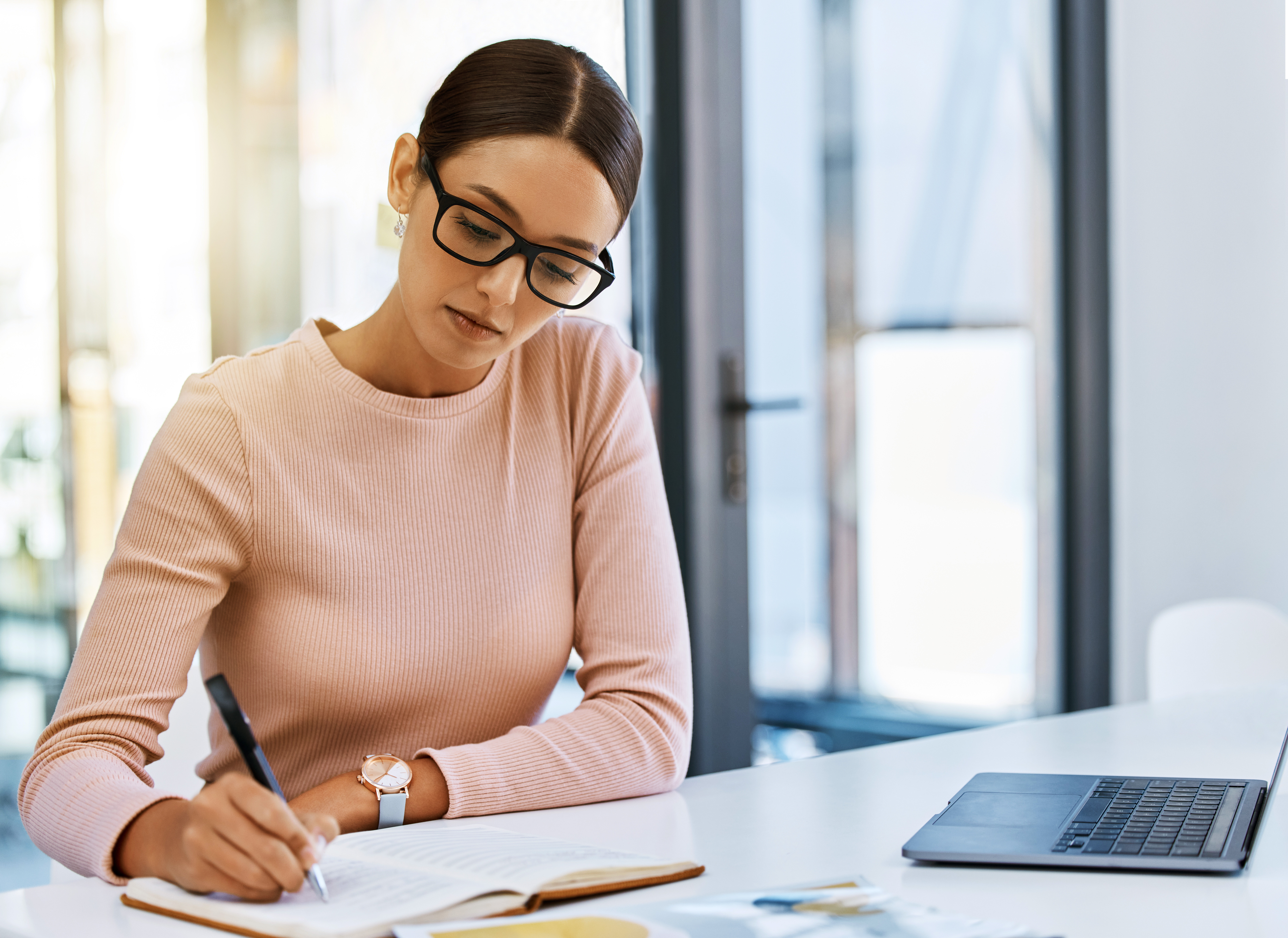 Woman with glasses writing in a notebook at a desk with a laptop.