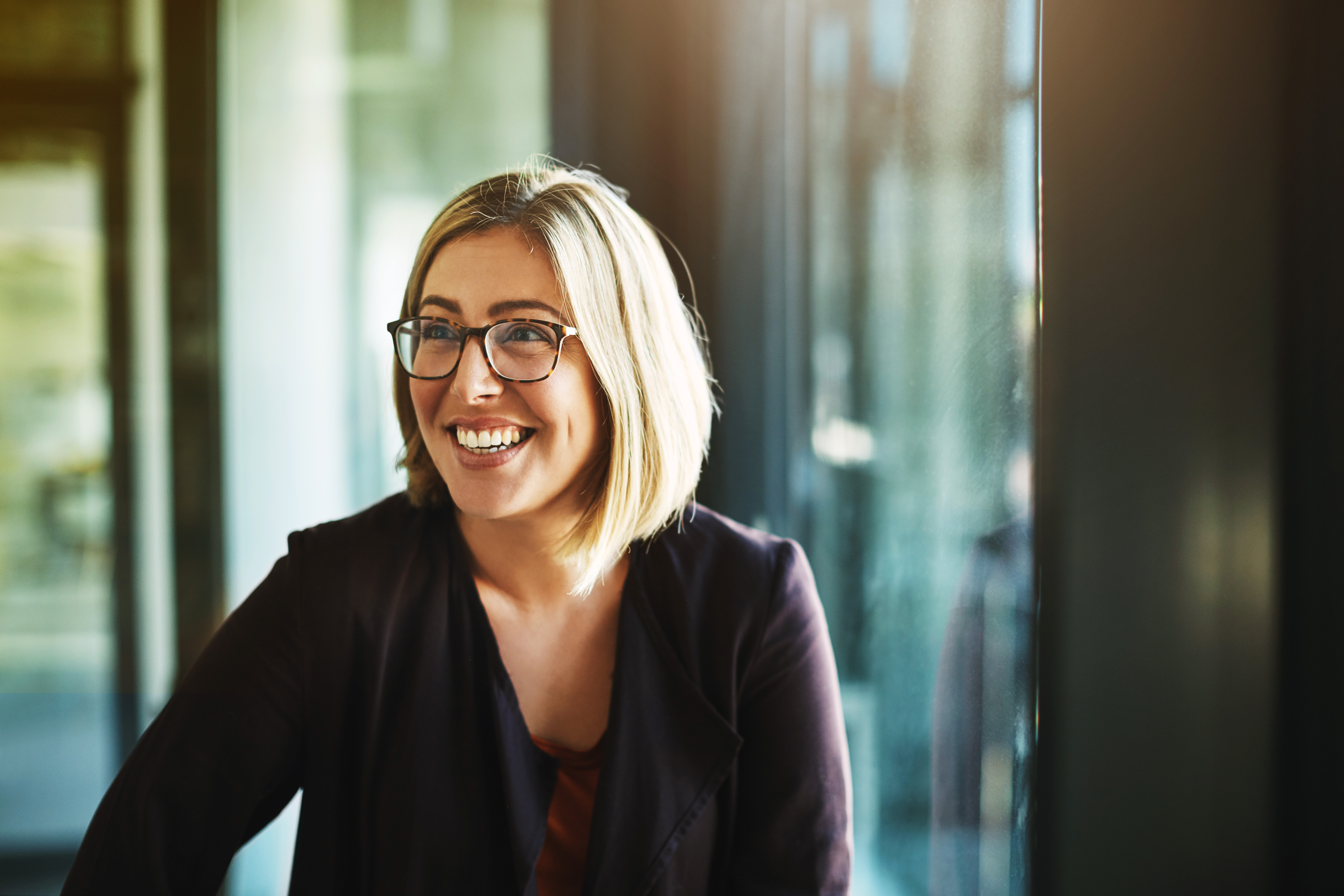 A smiling woman with glasses and a blazer stands by a window with a blurred background.