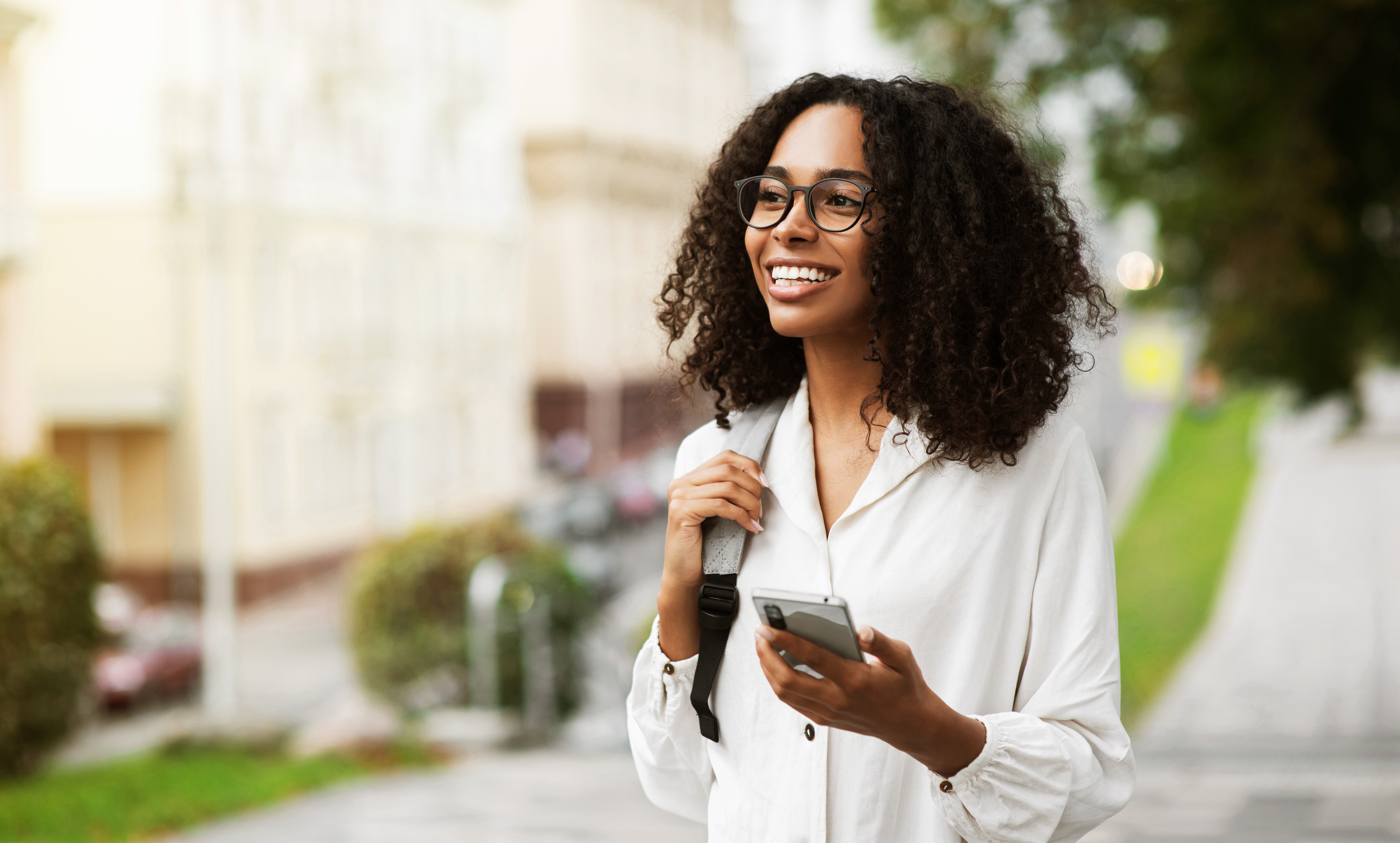 Smiling woman with curly hair wearing glasses holding a smartphone and backpack walking outdoors.