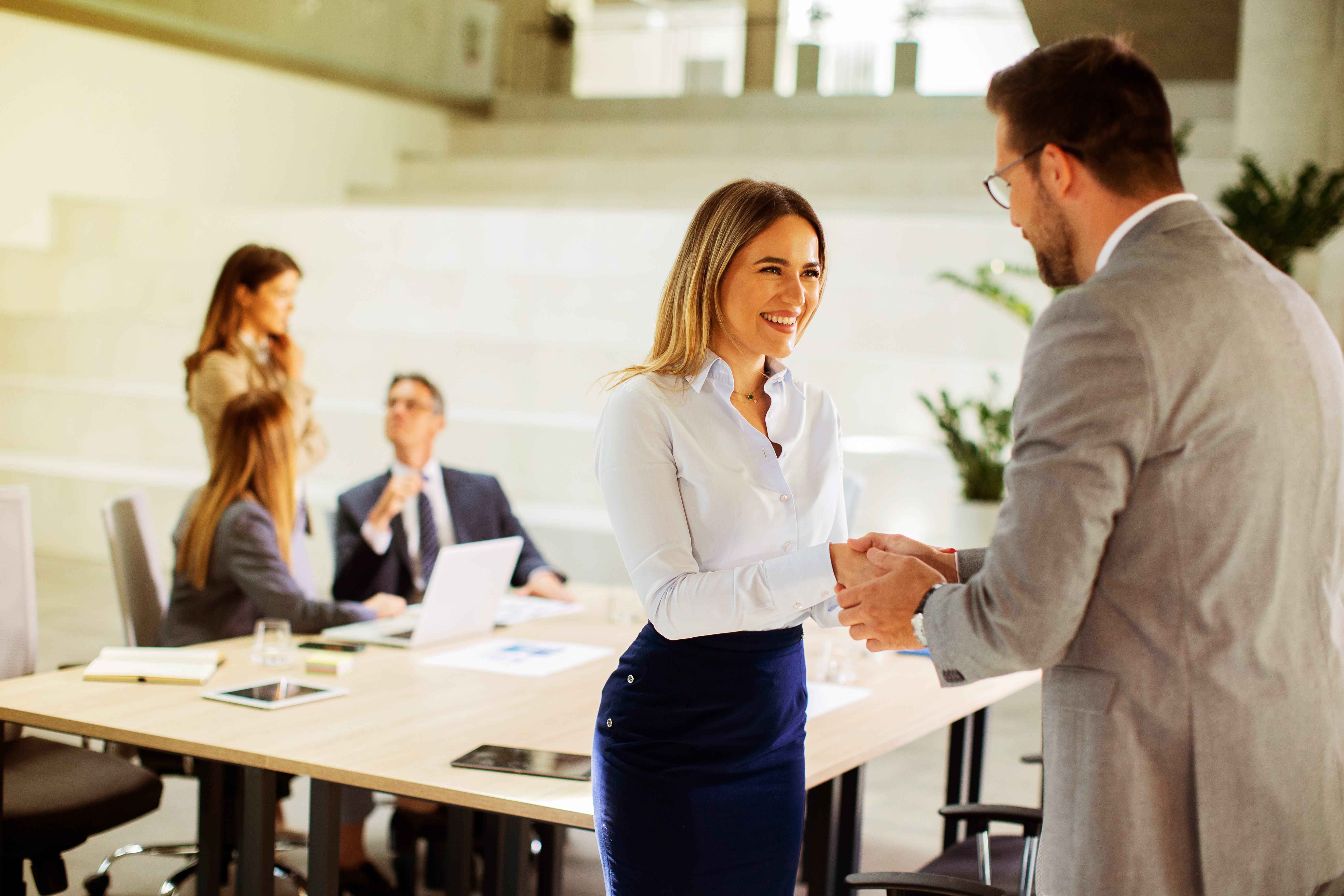 Businesspeople shaking hands in an office while two other colleagues discuss work in the background.