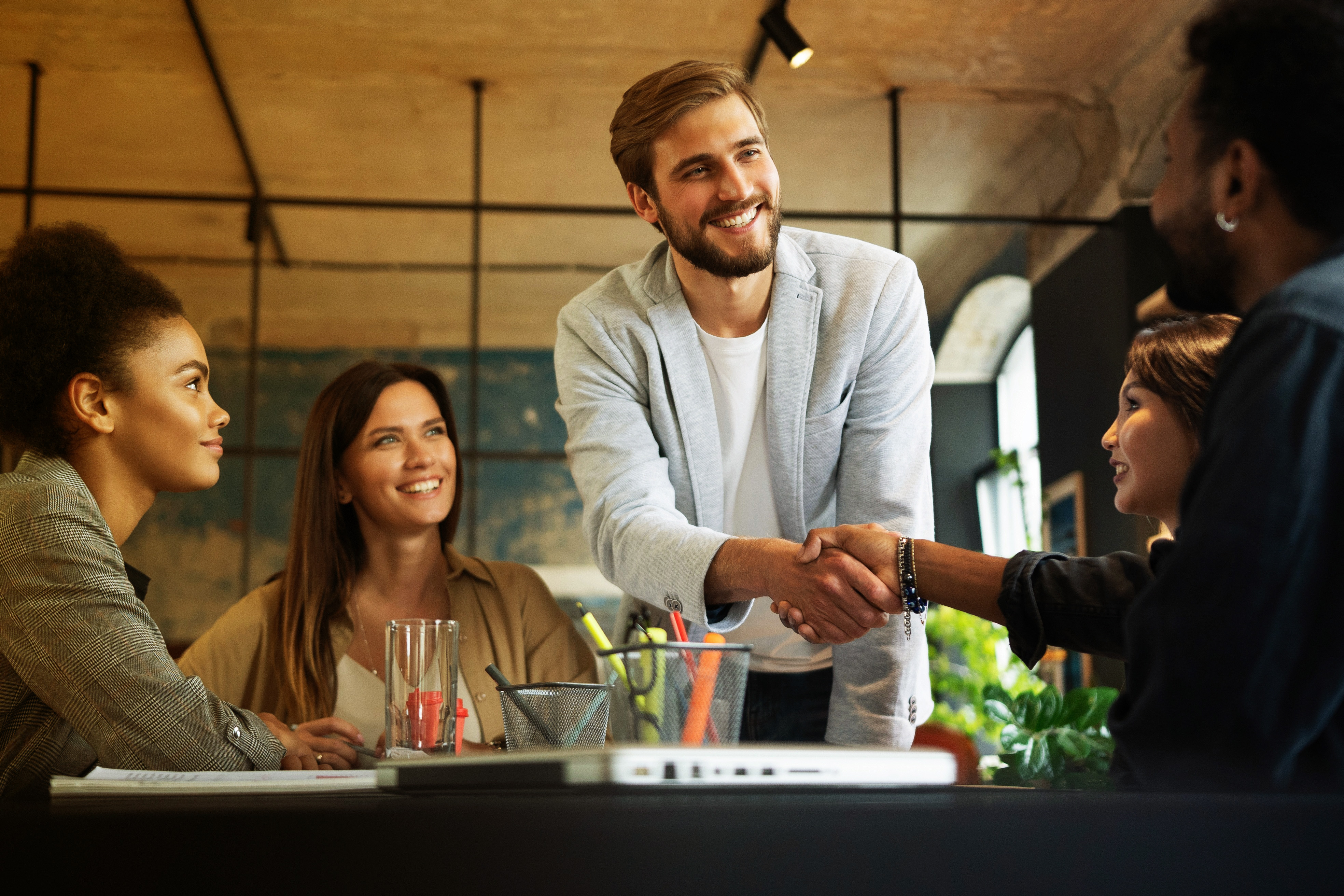 A man in a light blazer shakes hands with another man around a table with three smiling women.
