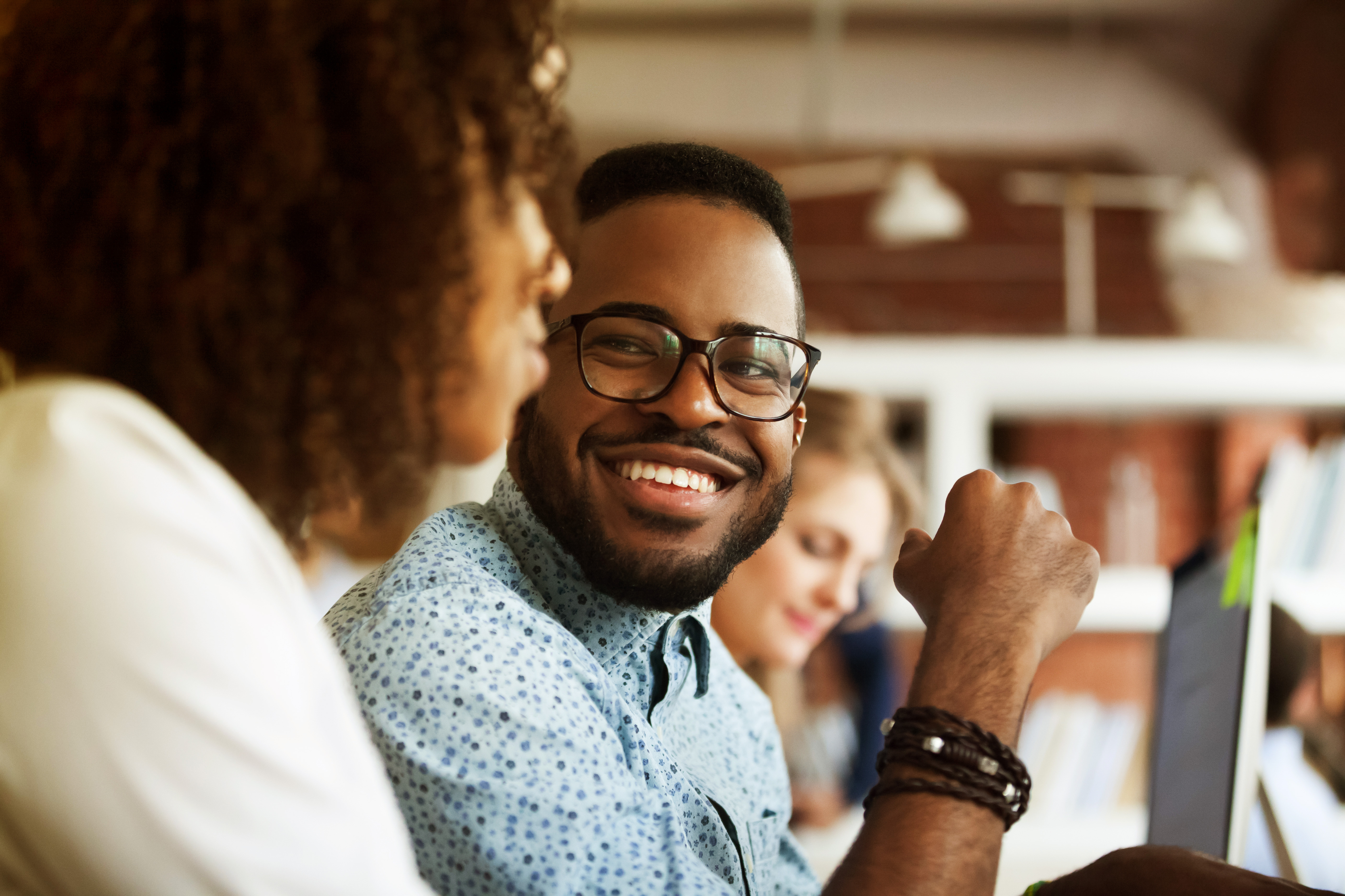 Two colleagues smiling and talking in an office setting with others working in the background.