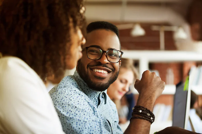 Smiling man with glasses speaking to a colleague, with others working in the background.
