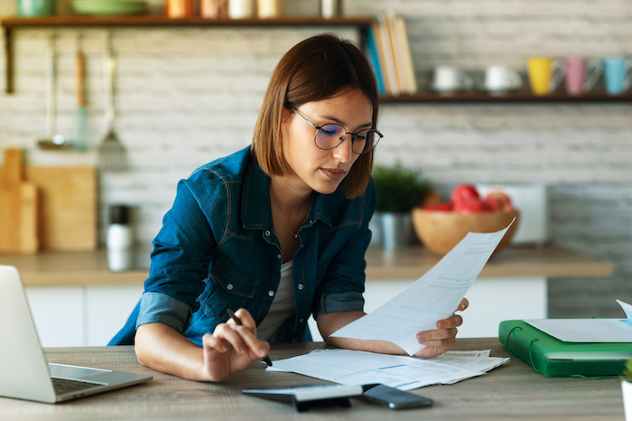 Woman with glasses reviewing papers and working on finances at a table with a laptop and calculator.