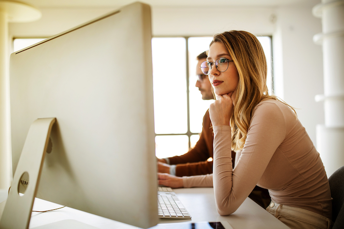Two professionals working at a desk with large monitors, one wearing glasses, deep in thought.