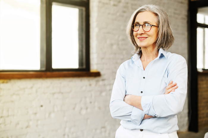 Older woman with glasses standing with arms crossed in an office wearing a light blue shirt.