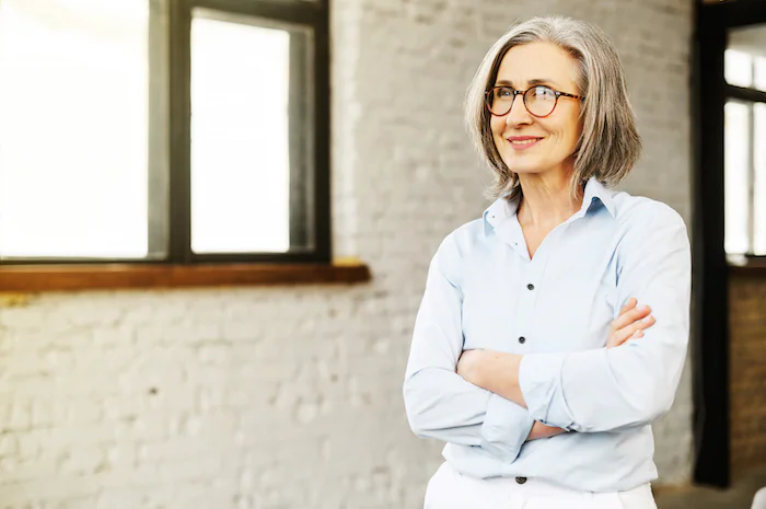 Older woman with glasses standing with arms crossed in an office wearing a light blue shirt.