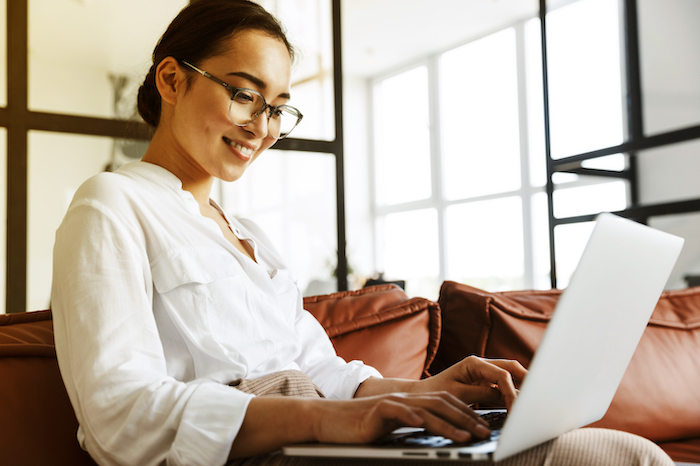 Woman wearing glasses and white shirt, typing on laptop in a bright room with large windows.