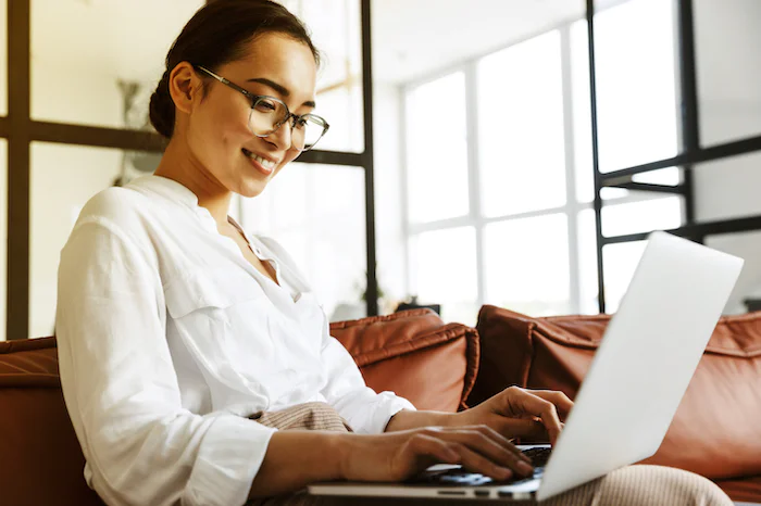 Smiling woman with glasses using a laptop on a brown leather couch in a bright room.