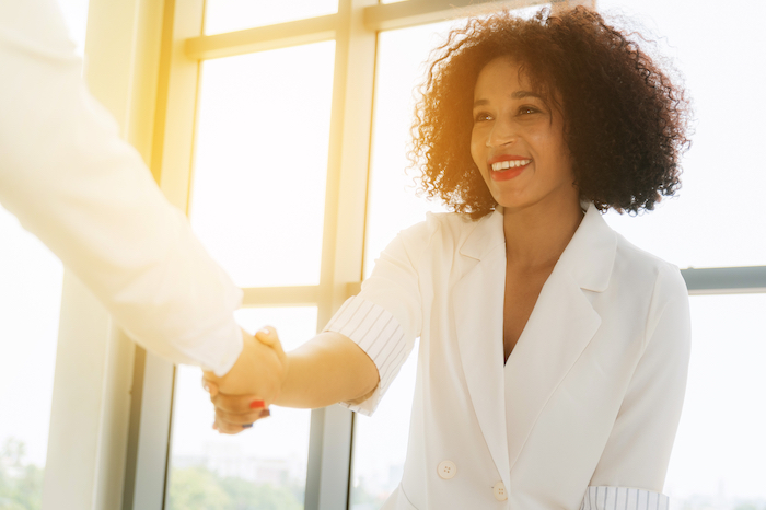 Smiling woman in white blazer shaking hands with another person in a bright office setting.