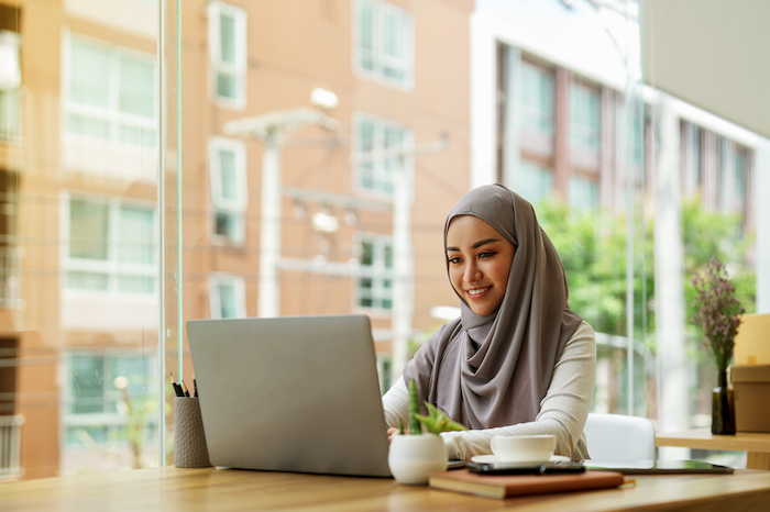 Woman in a hijab sitting at a cafe table using a laptop with buildings visible through the window.