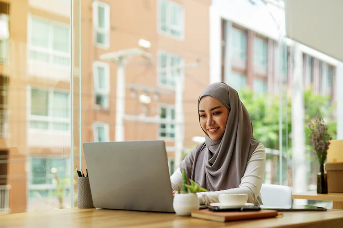 Woman in hijab working on a laptop at a desk near a large window in a modern office.