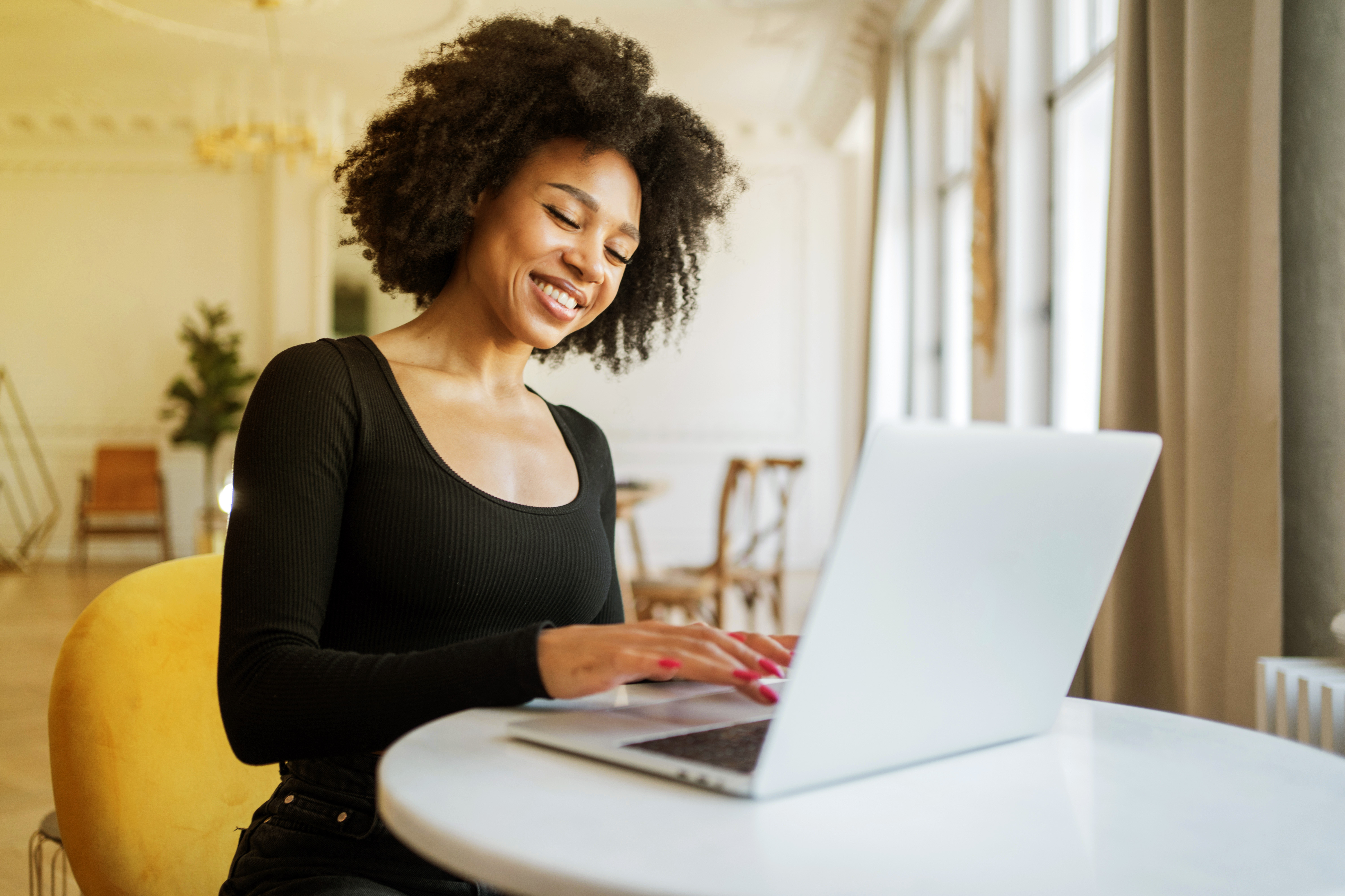 Woman with curly hair smiling and typing on a laptop at a round table