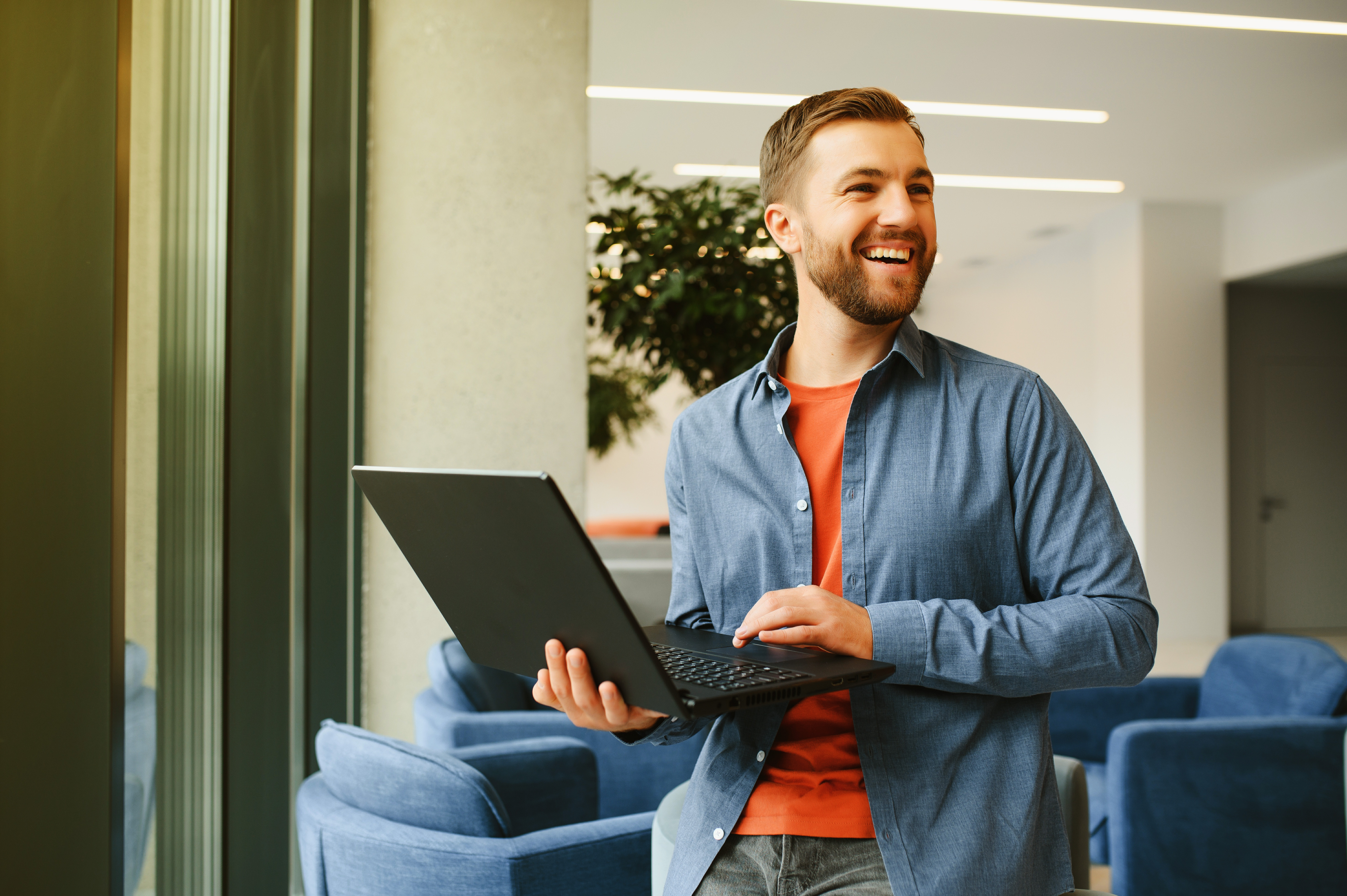 Man holding a laptop and smiling while standing in a modern office with blue chairs.