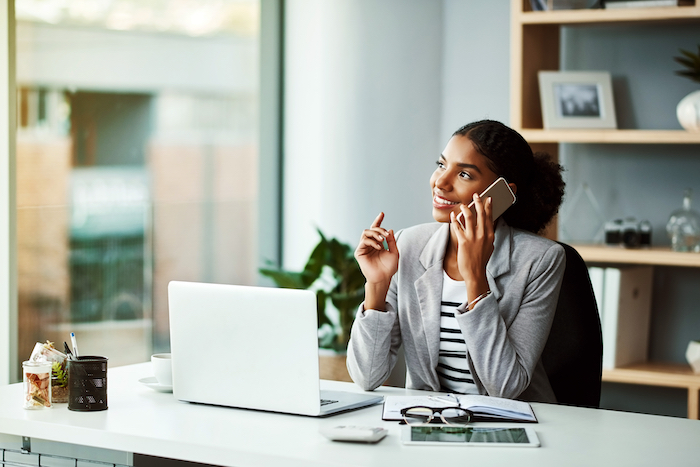Woman in a grey blazer talking on a phone while sitting at a desk with a laptop and documents.