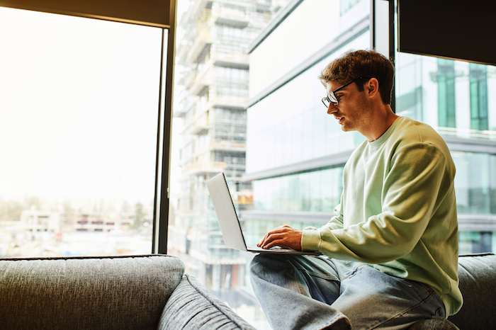 Man in glasses using a laptop while sitting on a couch near a window in a modern building.