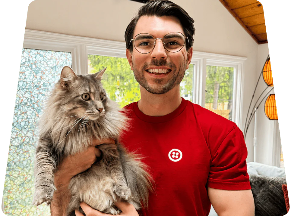 Smiling man wearing glasses holds a large fluffy cat indoors with large windows in the background.