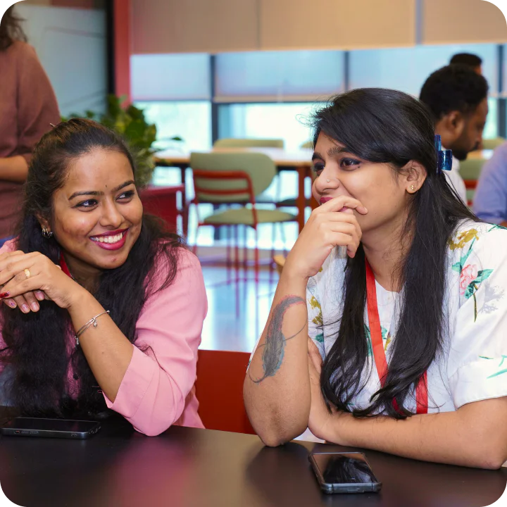Two women sitting at a table, smiling and talking in a bright, modern office setting.