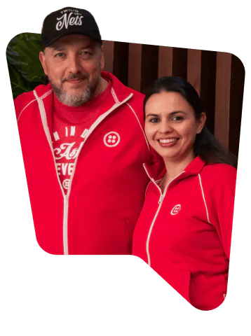 Two people in red jackets with matching logos, smiling in front of a wooden background.