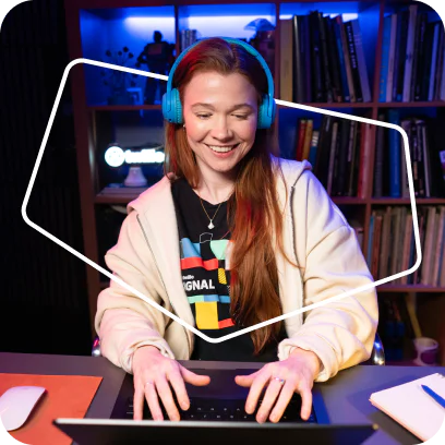 Woman wearing blue headphones and a colorful t-shirt, smiling while working on a laptop in a cozy room.