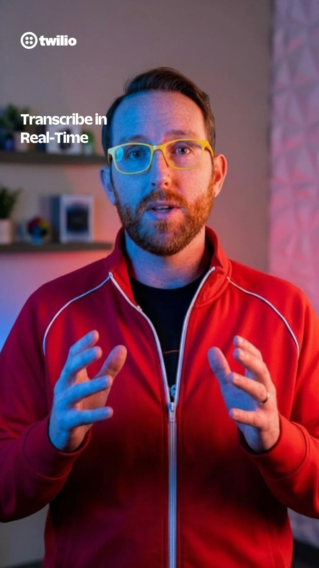 Man in red jacket and yellow glasses speaking with his hands in front of a wall and shelf.