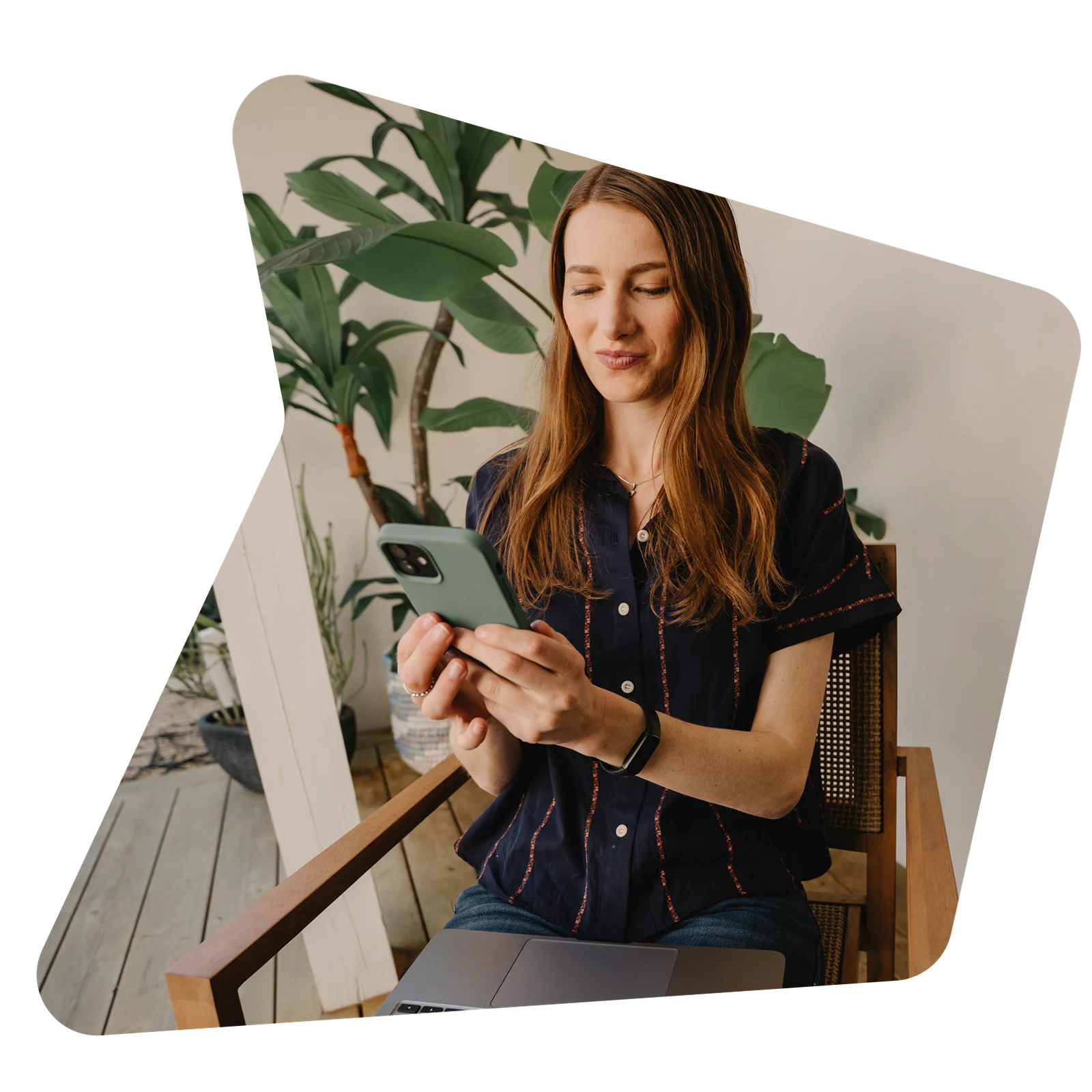 Woman sitting on a patio chair using a smartphone with a laptop on her lap, surrounded by plants.