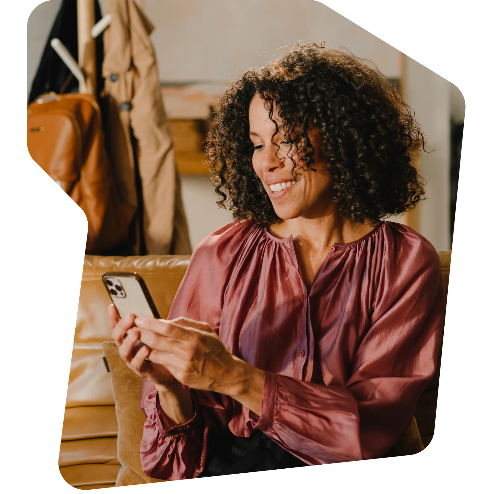 Smiling woman with curly hair wearing a pink blouse, looking at her phone while sitting on a leather couch.