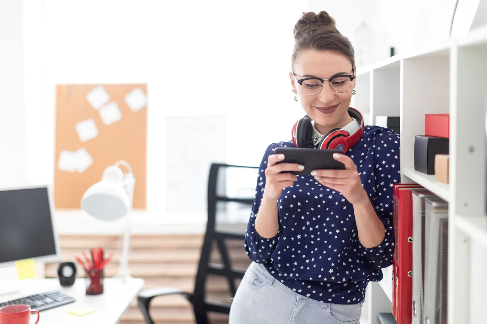 Young Professional Woman Working on Smartphone in Modern Office Woman with glasses and headphones, using a smartphone in a modern office, leaning against a shelf.