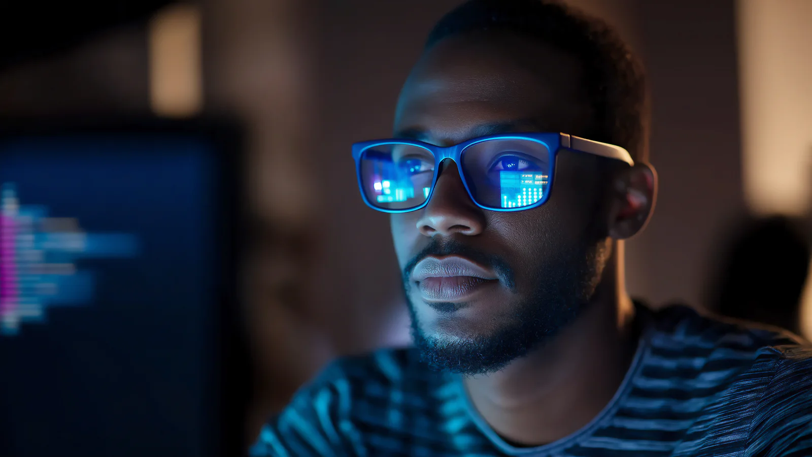 Close-up of a man wearing glasses, working on a computer at night. The scene is illuminated by the glow from the screen and the light reflecting off the lens. Man with glasses focused on computer screen displaying code at nighttime