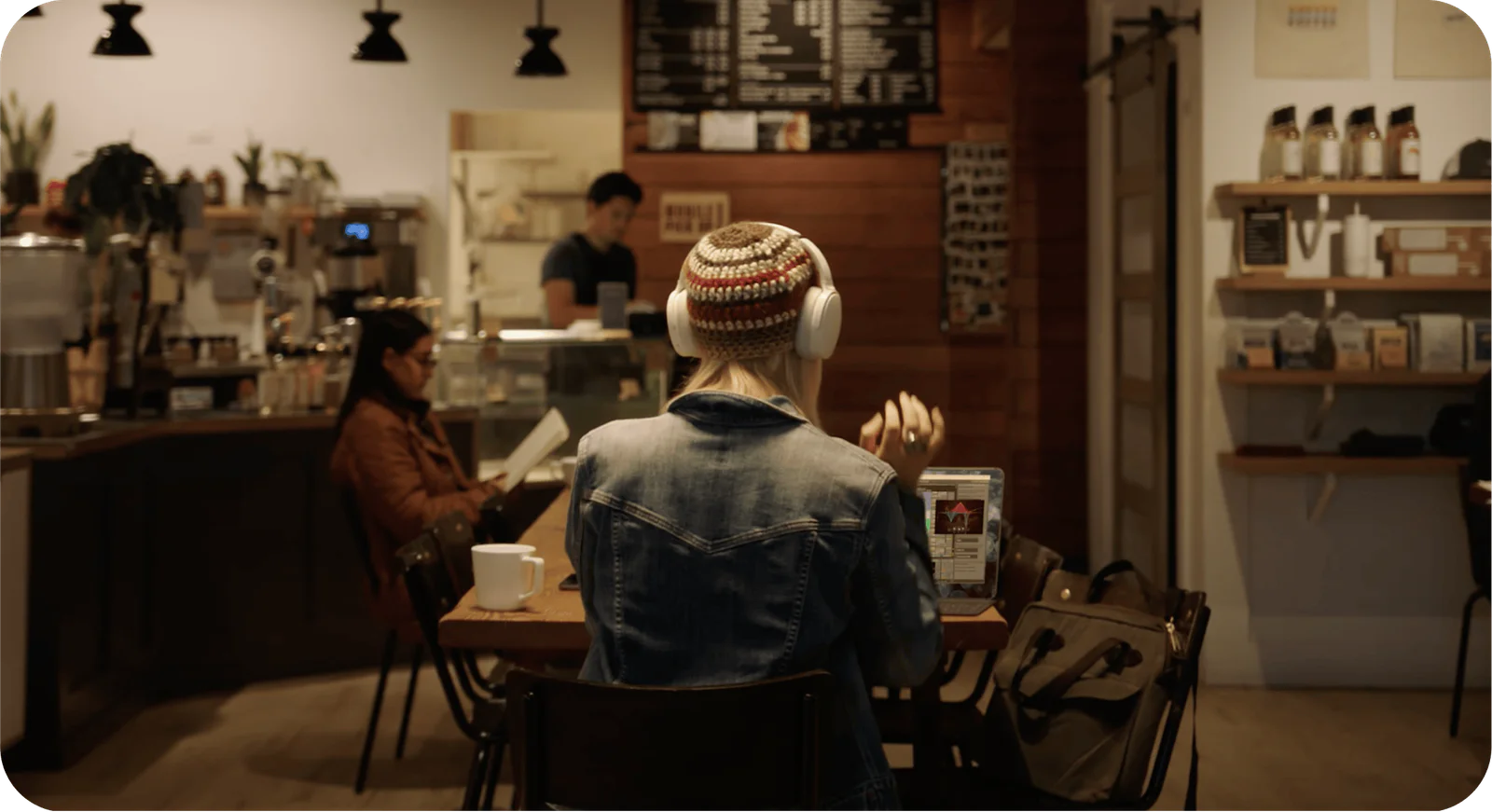 People sitting at a coffee shop, one wearing headphones and a beanie, with a barista in the background.