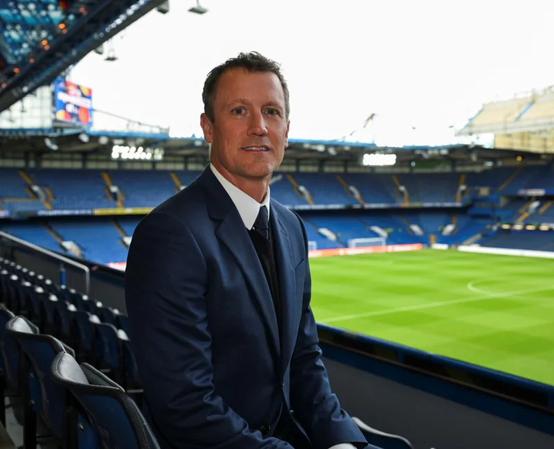 Man in a suit sitting in a stadium with empty seats and a soccer field in the background