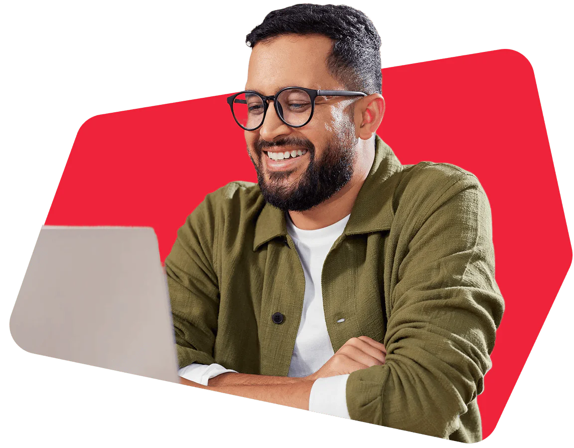 Man with glasses and beard smiling, using a laptop, red background.