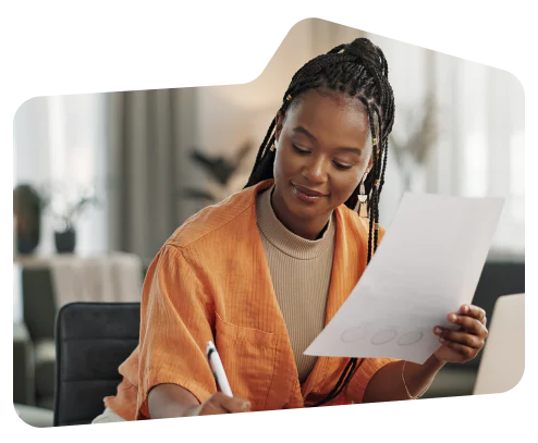 Woman in orange jacket holding a paper and pen, sitting at a desk with a thoughtful expression.