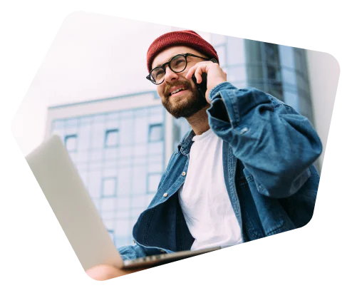 Man in red beanie and glasses using a laptop and talking on a phone outside a modern building.