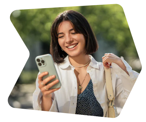Woman with short hair smiling at her smartphone while holding a shoulder bag outdoors.