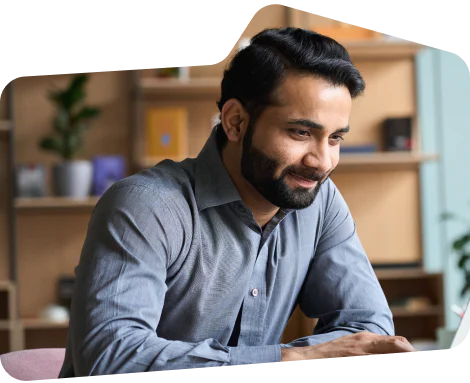 Smiling man in gray shirt sitting at desk using a laptop in an office setting