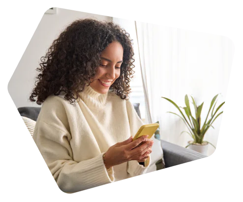 Woman with curly hair wearing a white sweater smiling and looking at her smartphone.
