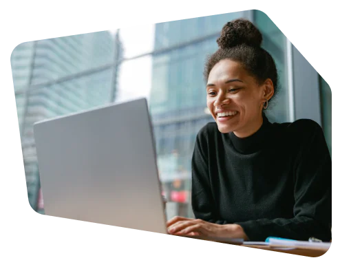 Woman smiling and typing on a laptop while seated by a window in a modern office setting