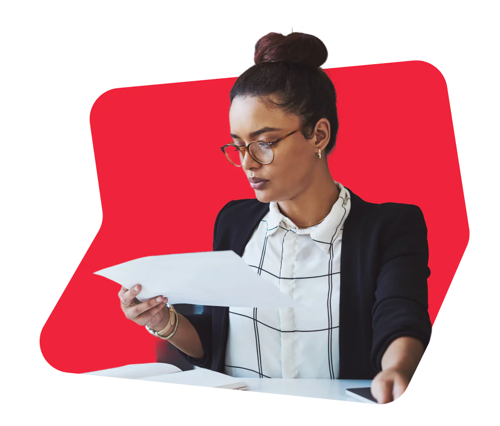 Woman in glasses and professional attire reading documents at a desk with a red background.