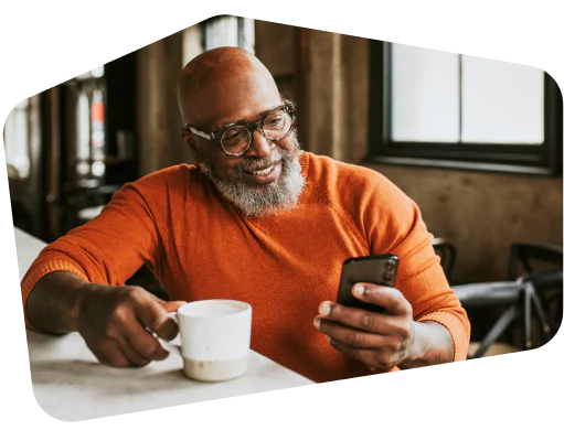 A man in an orange sweater looking at his smartphone and holding a cup of coffee