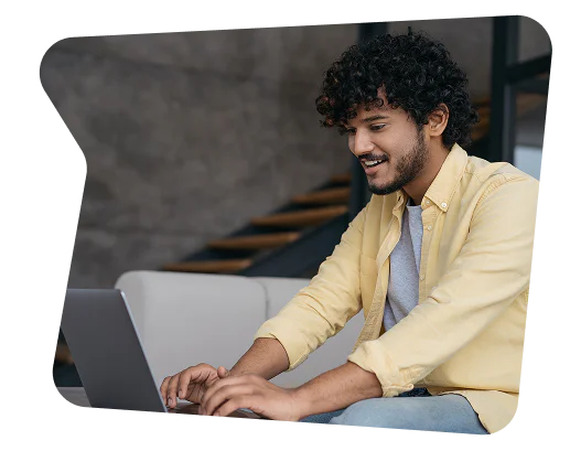 Young man with curly hair and a yellow shirt using a laptop inside a modern room.
