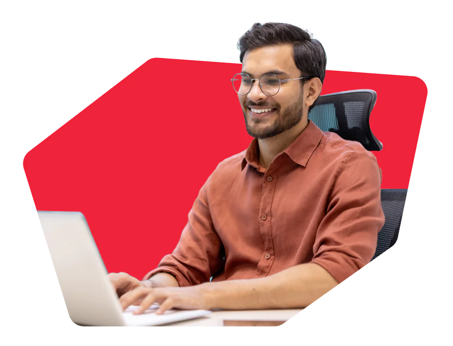 Smiling man with glasses in a brown shirt working on a laptop against a red background.