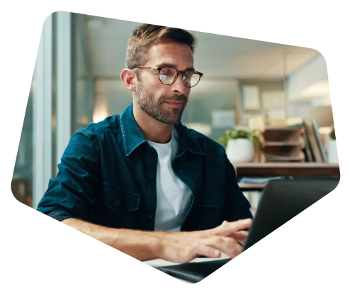 Man with glasses and beard typing on laptop in a modern office with books and plants in the background.