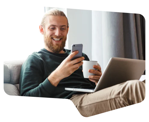 Man smiling, using smartphone, holding a coffee cup, and sitting with a laptop on his lap.